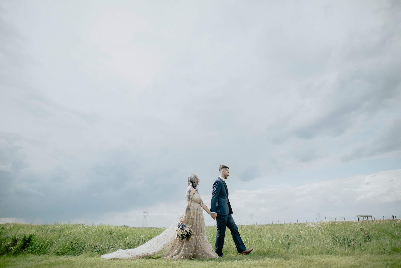 A bride and groom walking in a field under a cloudy sky.