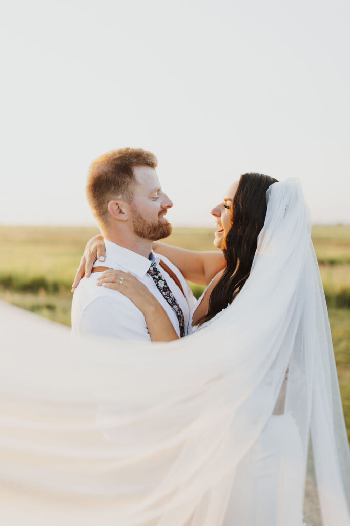 A bride and groom standing in a field with their veil blowing in the wind at the Gathered an outdoor wedding venue near Calgary, Ab.