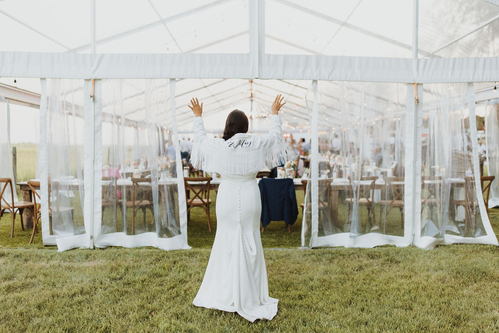 A bride in a white dress and jacket saying wifey standing in front of a tent.