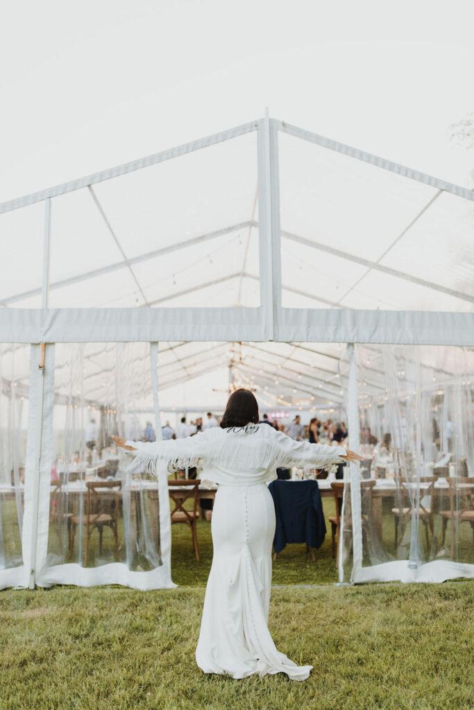 A bride standing in front of a tent wearing a wifey jacket. Enchanting Outdoor Dinner Party.