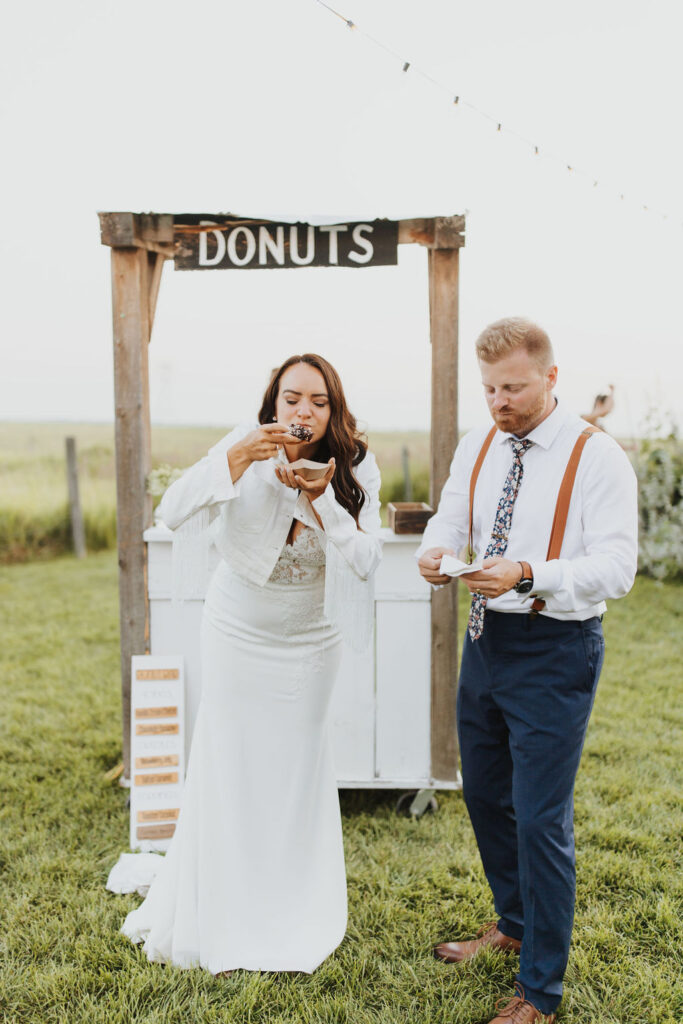 A bride and groom eating donuts in front of a donut stand.