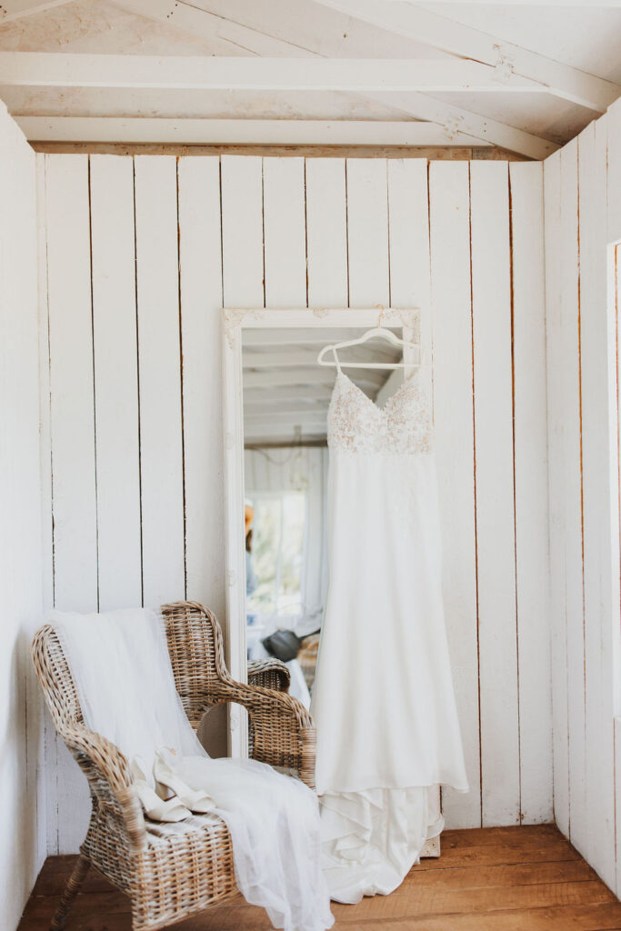 A wedding dress hangs on a wicker chair in a room with white walls.
