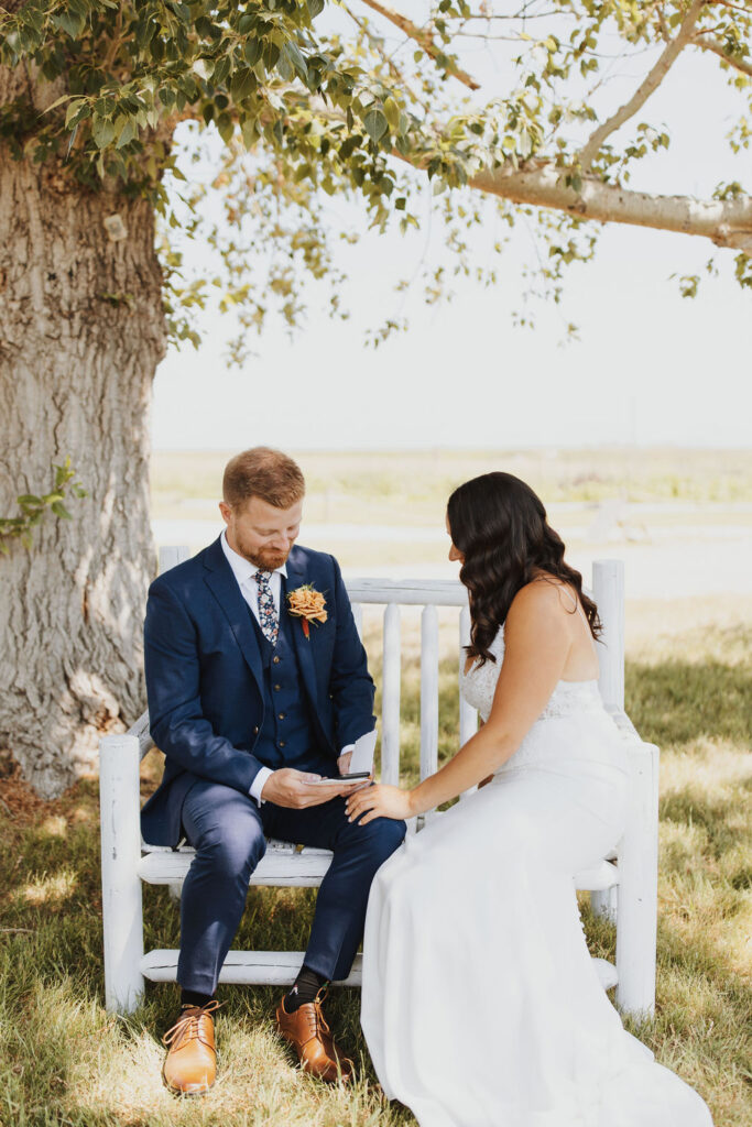 A bride and groom sitting on a white bench in a field.