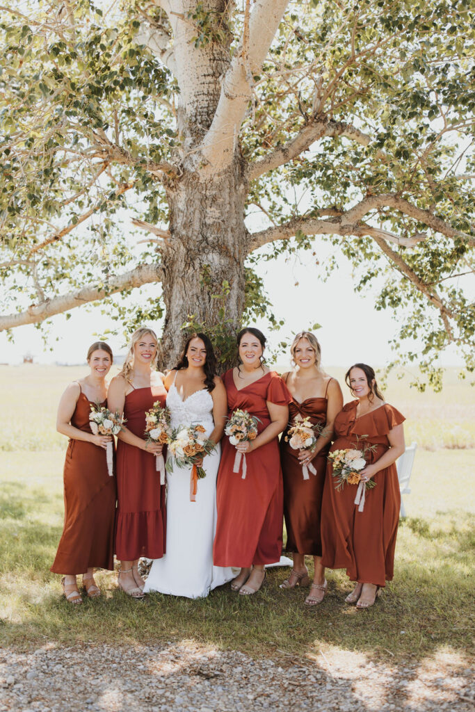 Bridesmaids in burgundy dresses standing in front of a tree.