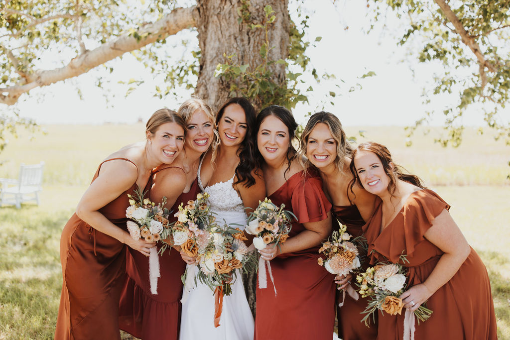 Bridesmaids in burgundy dresses pose for a photo in front of a tree.
