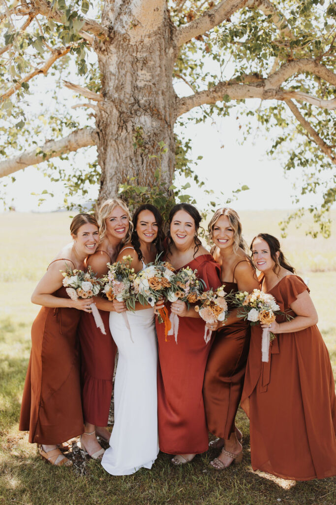 Bridesmaids in burgundy dresses pose in front of a tree.