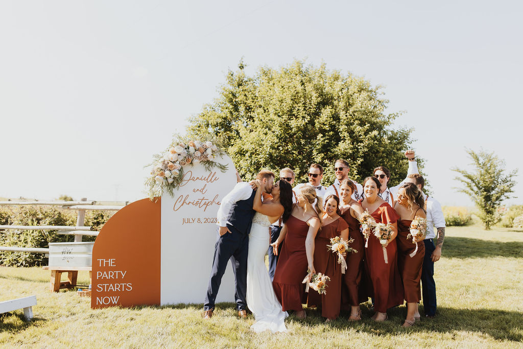 A group of bridesmaids and groomsmen pose in front of an orange sign. 