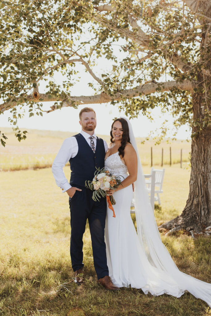 A bride and groom standing under a tree in a field.