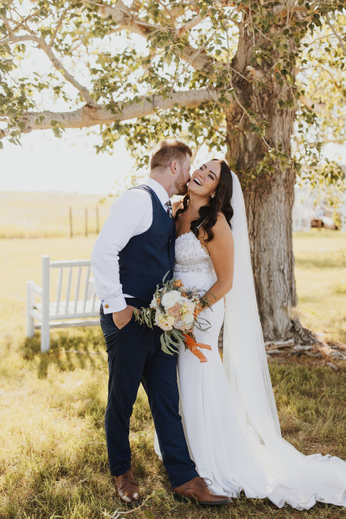 A bride and groom kiss in front of a tree.