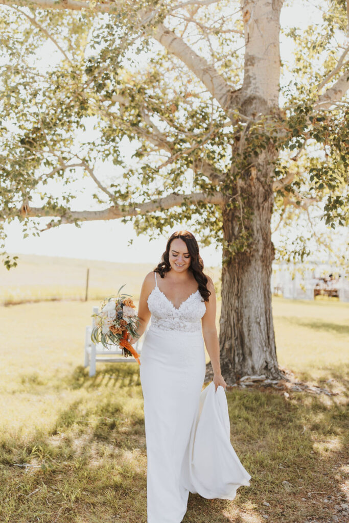 A bride in a white dress walks under a tree.