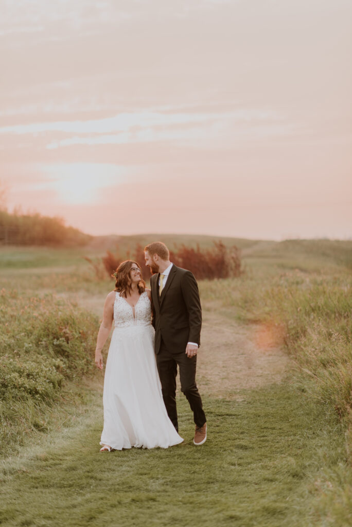 A bride and groom walking down a grassy path at sunset.