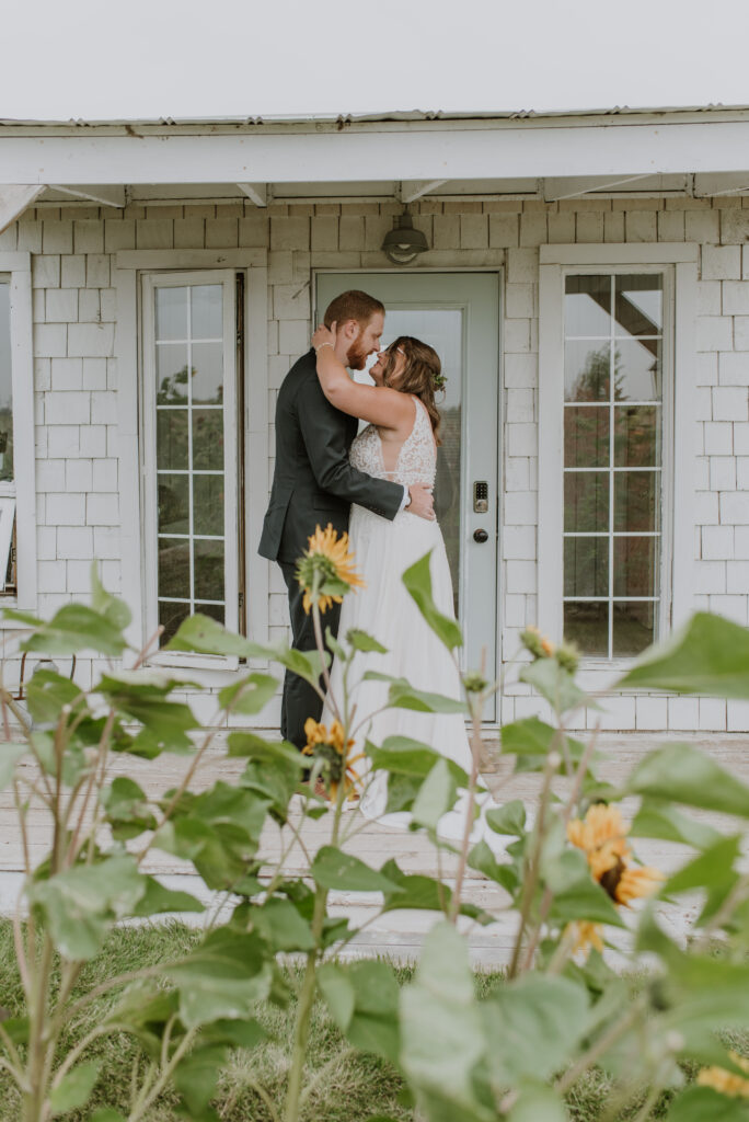 A bride and groom kiss in front of the Gathered cottage with sunflowers in the background.