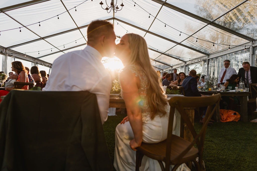 A bride and groom kissing in a tent at sunset.