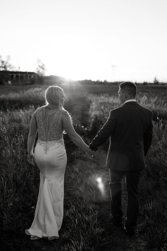 A bride and groom walking through a field at sunset.
