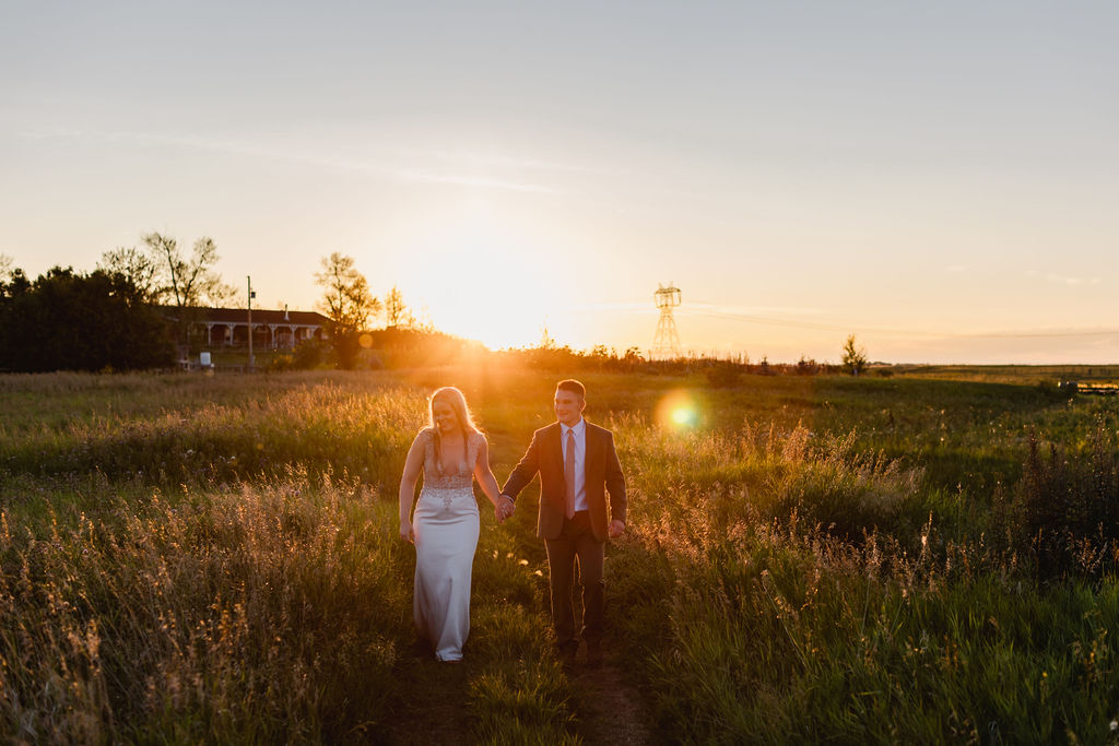A bride and groom walking through a field at sunset.