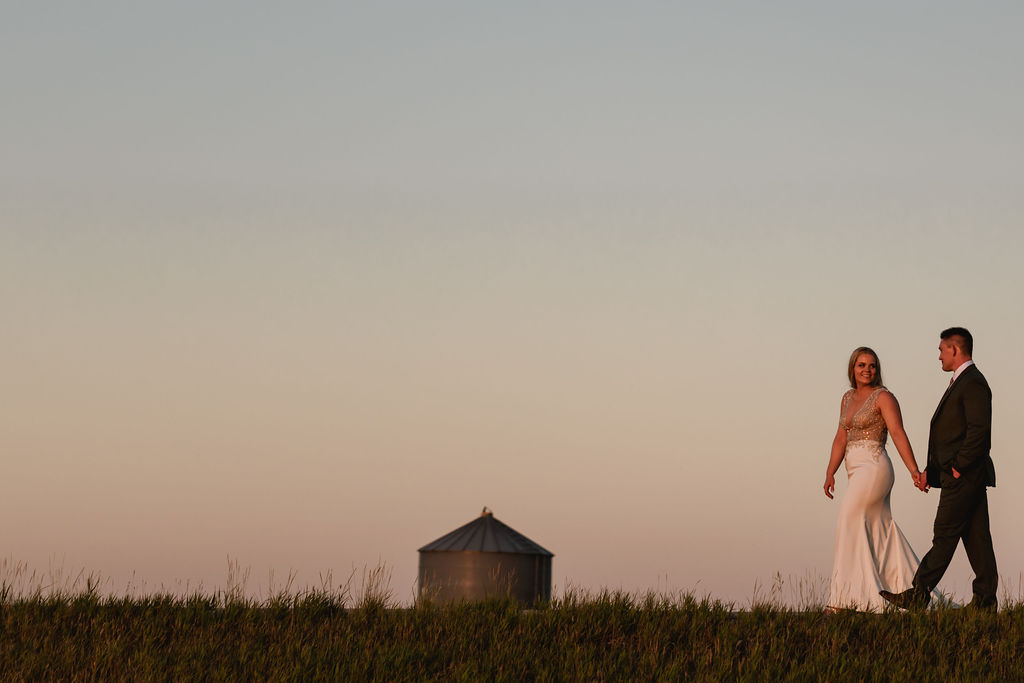 A bride and groom walking in a field at sunset.