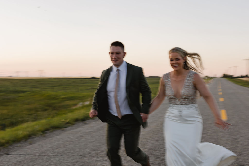 A bride and groom running down a road at sunset.