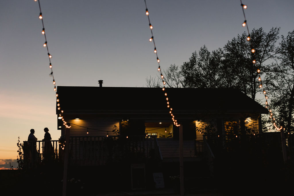 A couple sits on the porch of a house at dusk.