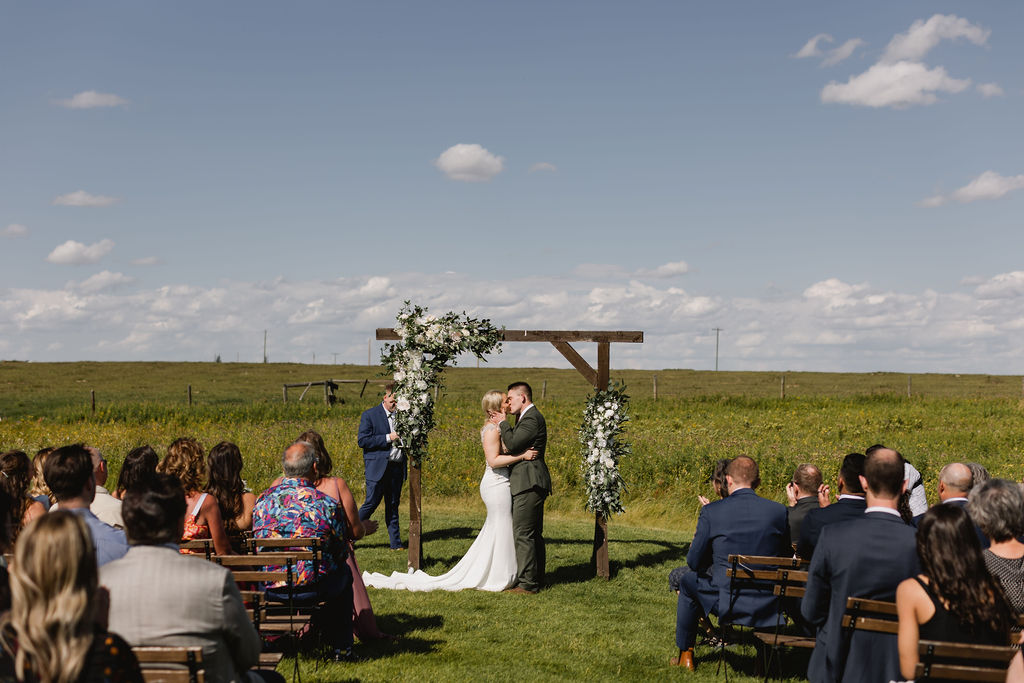 A bride and groom kiss during their wedding ceremony in a field.