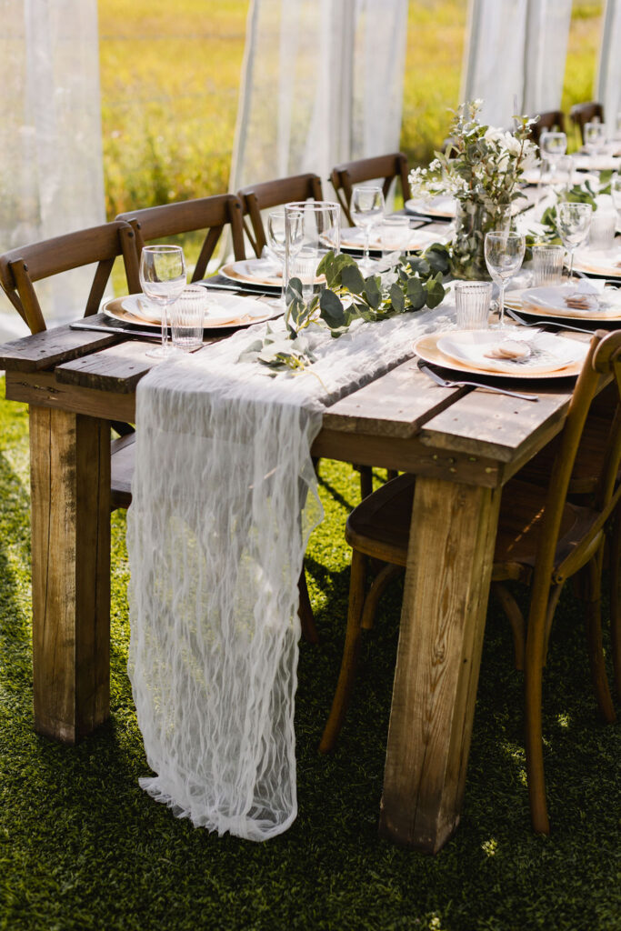 A table set up in a tent with white gauze table runners, greenery and diy wedding florals on wood harvest tables. 