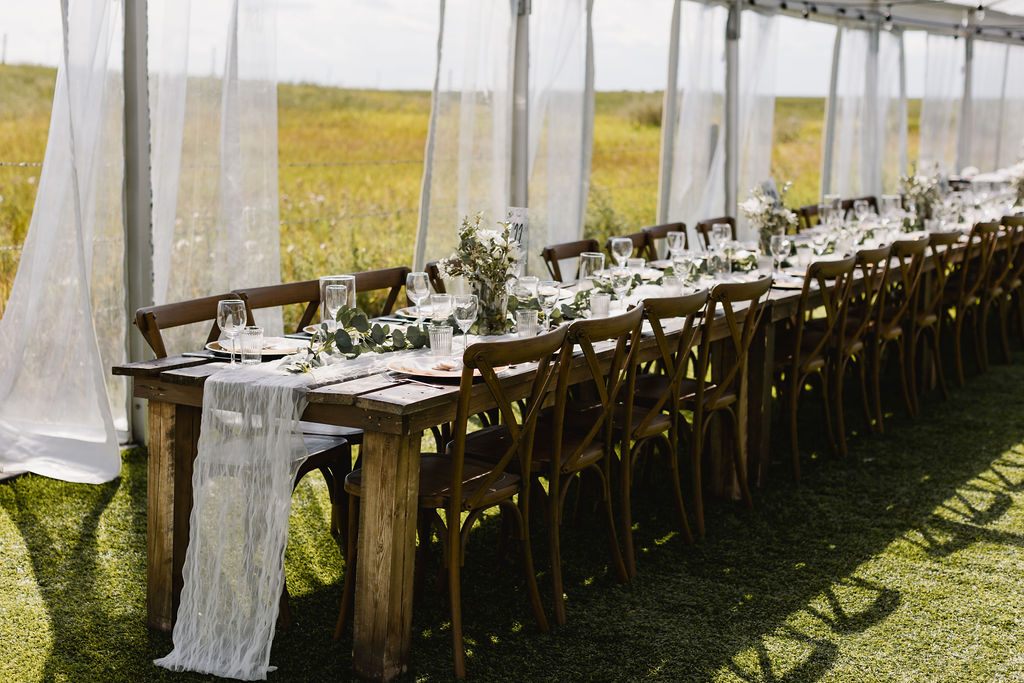 A long table set up in a grassy field. Wood harvest tables, white gauze table runners and diy florals 
