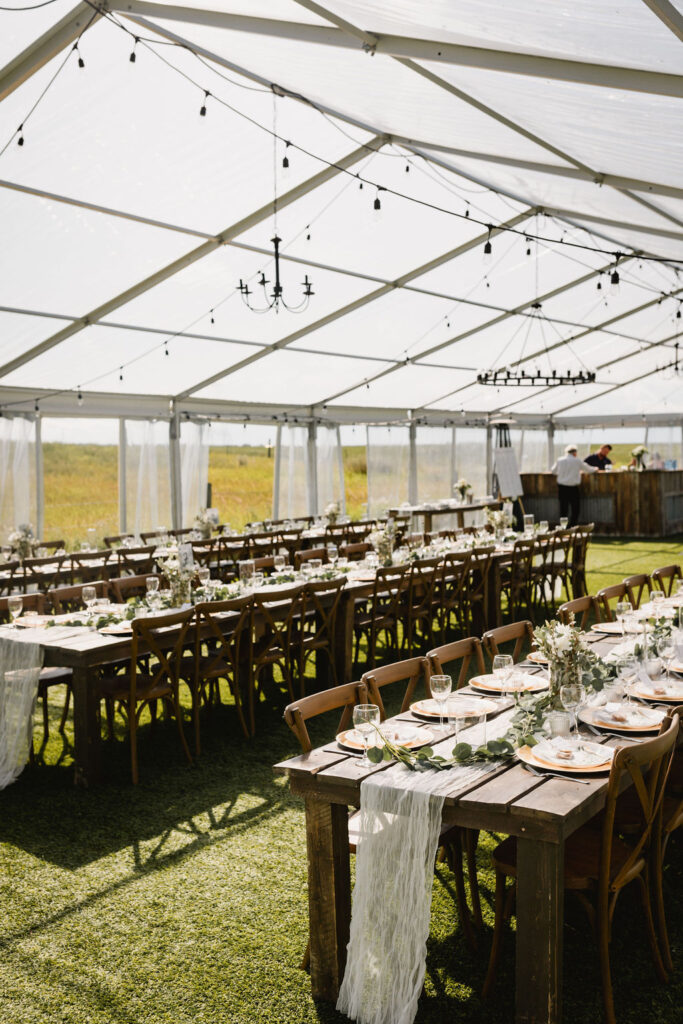 A clear tent set up for an outdoor wedding reception with wood harvest tables, hanging black chandeliers and globe lights. DIY wedding florals and centrepieces. 
