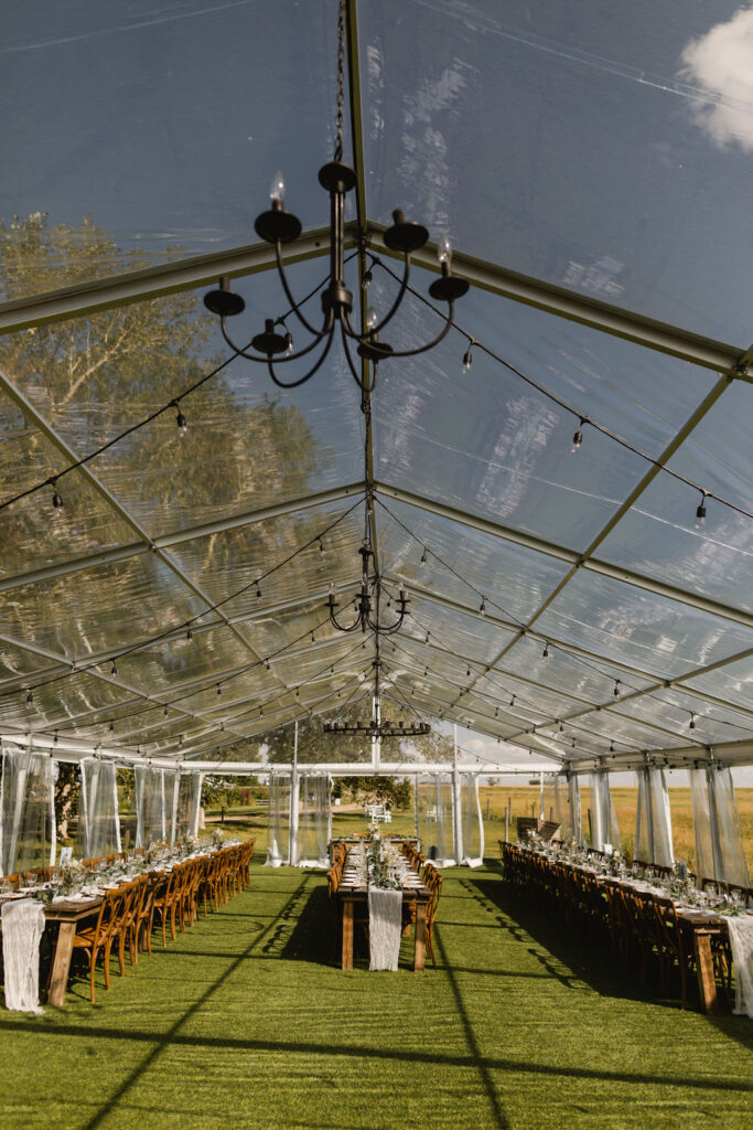 A clear tent set up for a wedding reception with wood harvest tables, hanging globe lights and DIY florals.