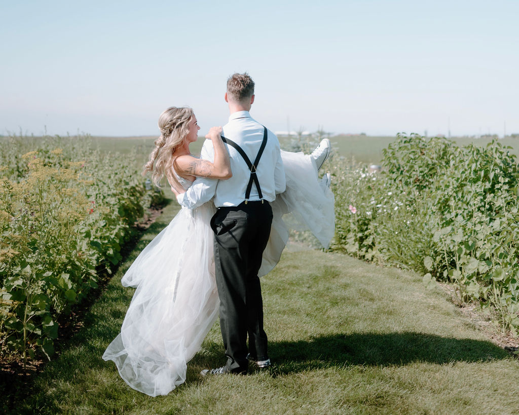 Bride and groom celebrating by walking through a field, with the groom carrying the bride's dress.