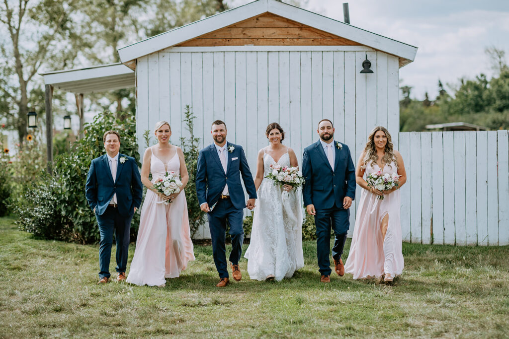 A wedding party posing outside with the bride and groom in the center, flanked by groomsmen in blue suits and bridesmaids in pastel dresses.
