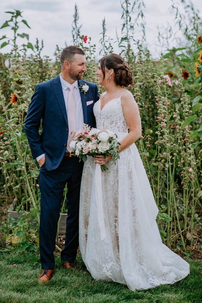 A bride and groom smiling at each other in a garden with sunflowers in the background.