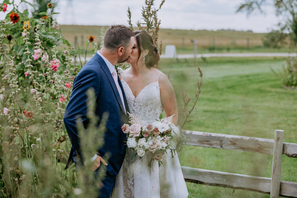 Bride and groom sharing a kiss beside a garden fence while holding a bouquet.