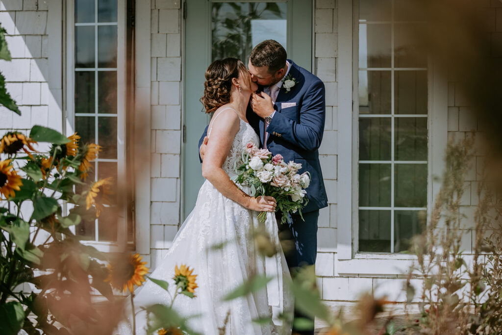 A couple in wedding attire sharing a kiss outside a white window framed by sunflowers.