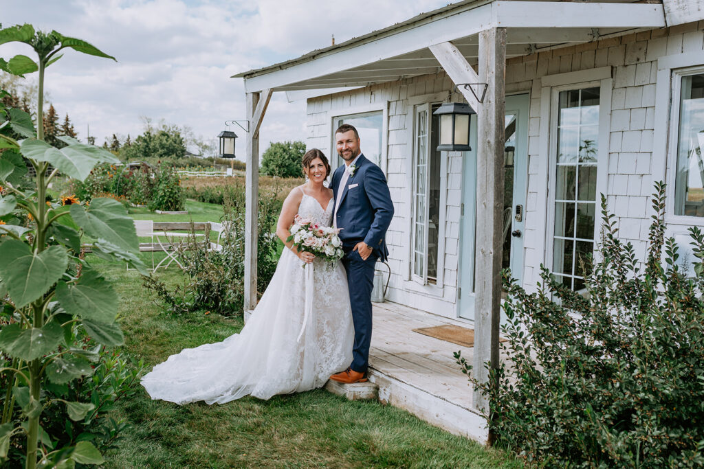 A bride and groom smiling by the doorway of a rustic white cabin with greenery around.