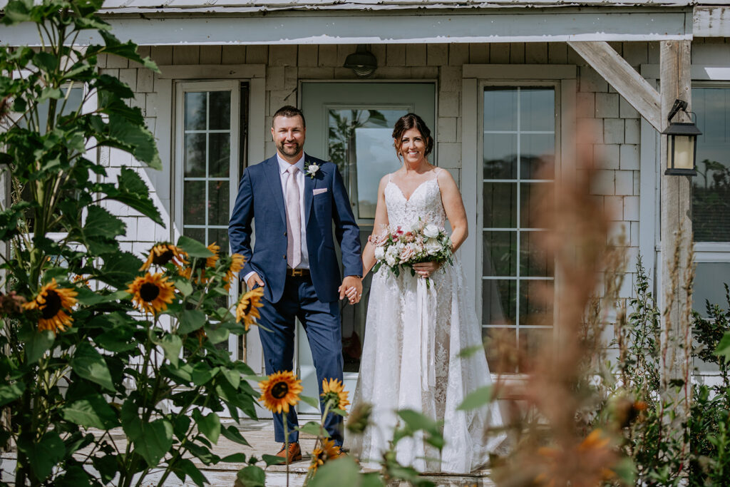 A bride and groom holding hands in front of a white house, surrounded by greenery and sunflowers.