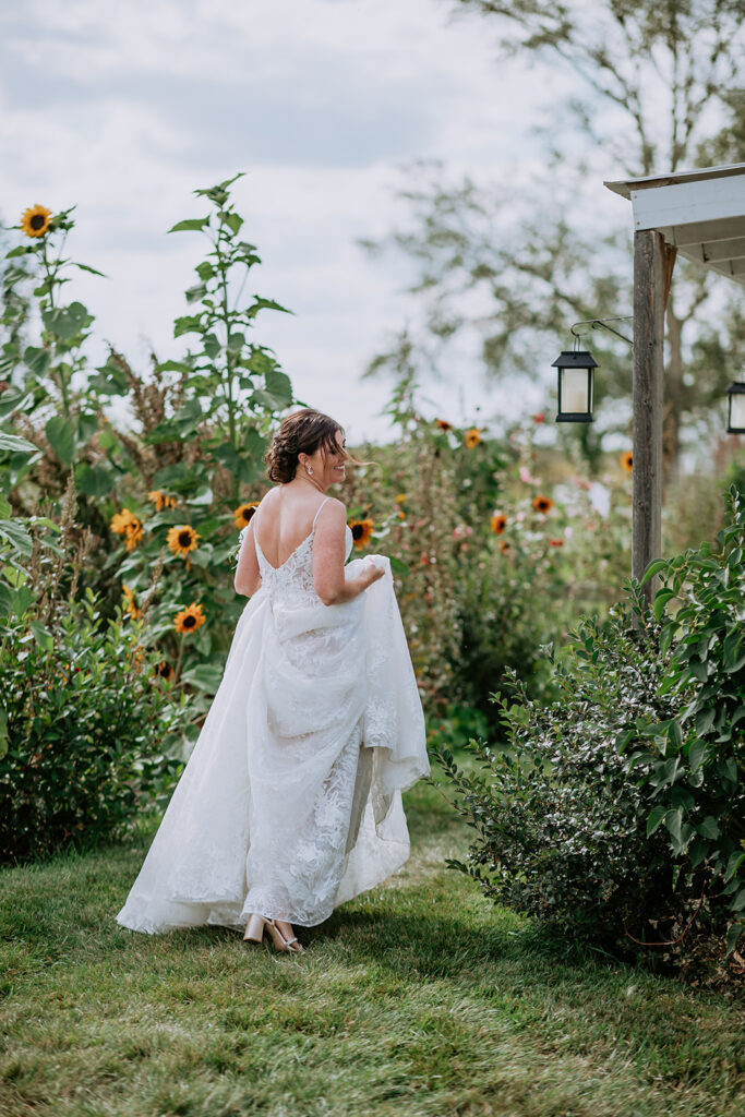 A bride in a white dress holding up her skirt as she walks through a garden with sunflowers.