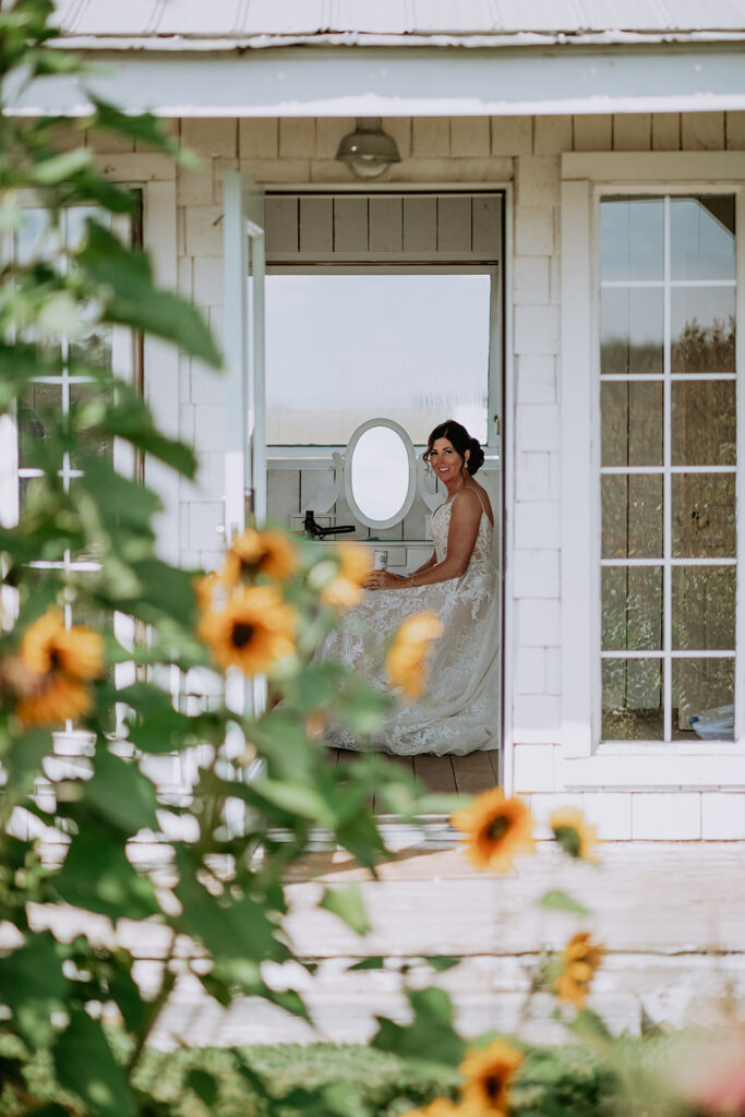 A bride in a white dress sitting by a makeup mirror in a doorway, framed by sunflowers.