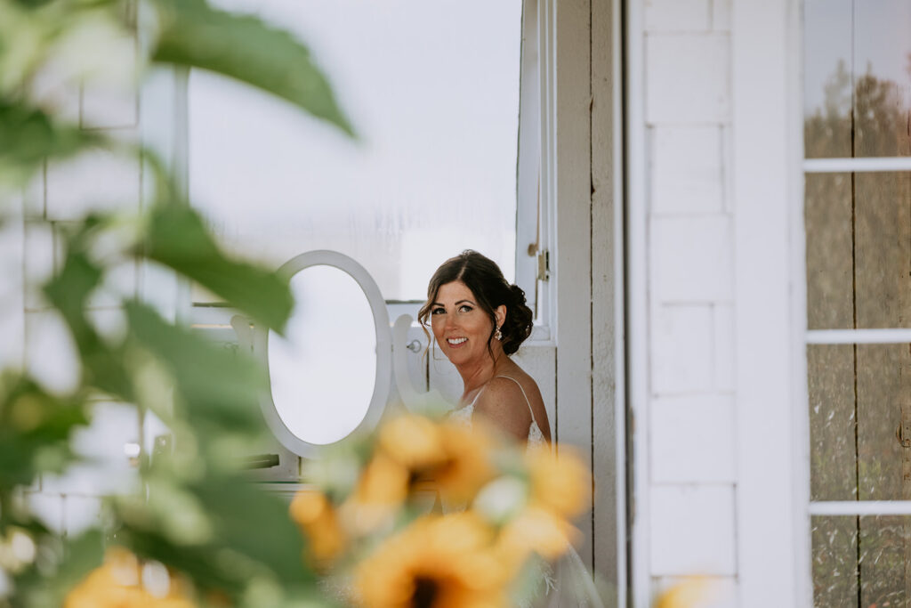 A woman smiling over her shoulder by a window, framed by greenery and sunflowers.