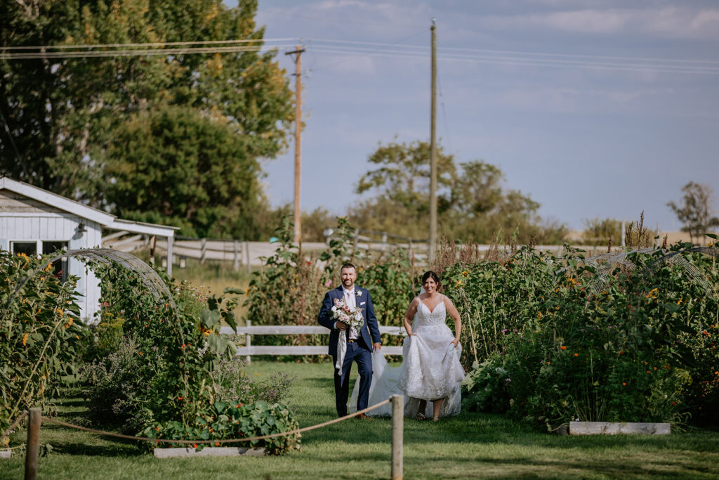 A bride and groom walking through a garden with the groom leading the way and the bride following, both smiling and holding hands.