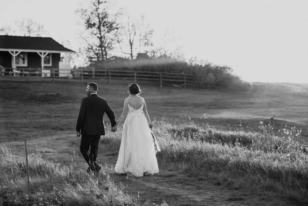 Bride and groom walking hand in hand through a field at sunset.