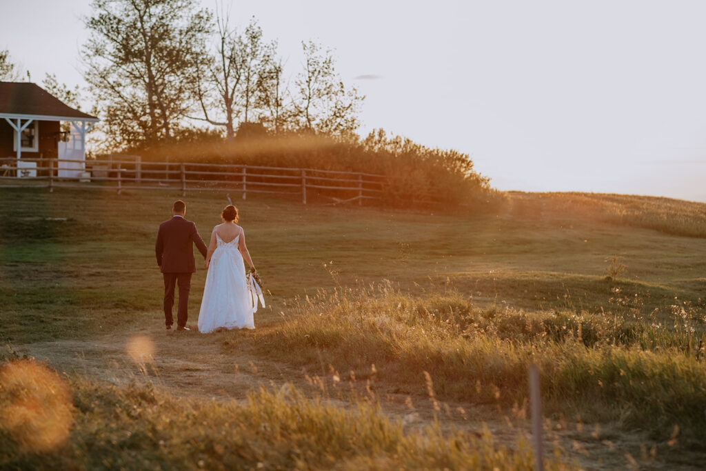 A couple in wedding attire walking hand in hand through a field at sunset.