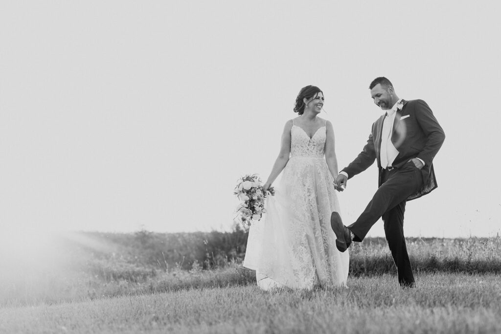 A black and white photo of a bride and groom walking through a field, smiling and holding hands, with the groom playfully lifting one leg.