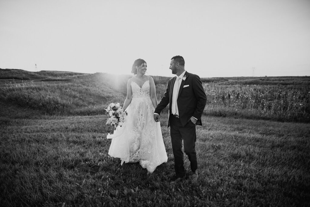 A bride and groom walking hand in hand in a field, with the sun casting a gentle glow beside them.