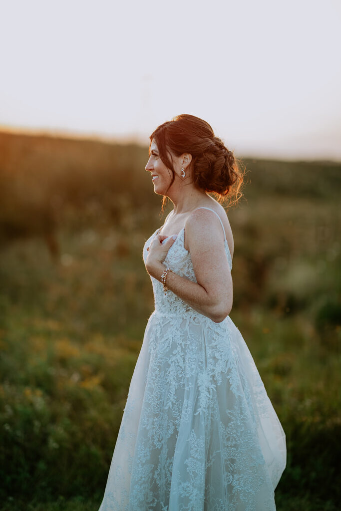 Bride in lace wedding gown smiling outdoors at sunset.
