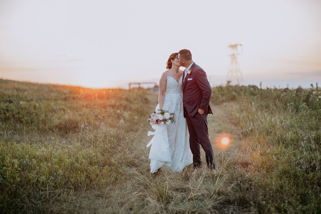 A couple on their wedding day sharing a kiss in a grassy field at sunset.