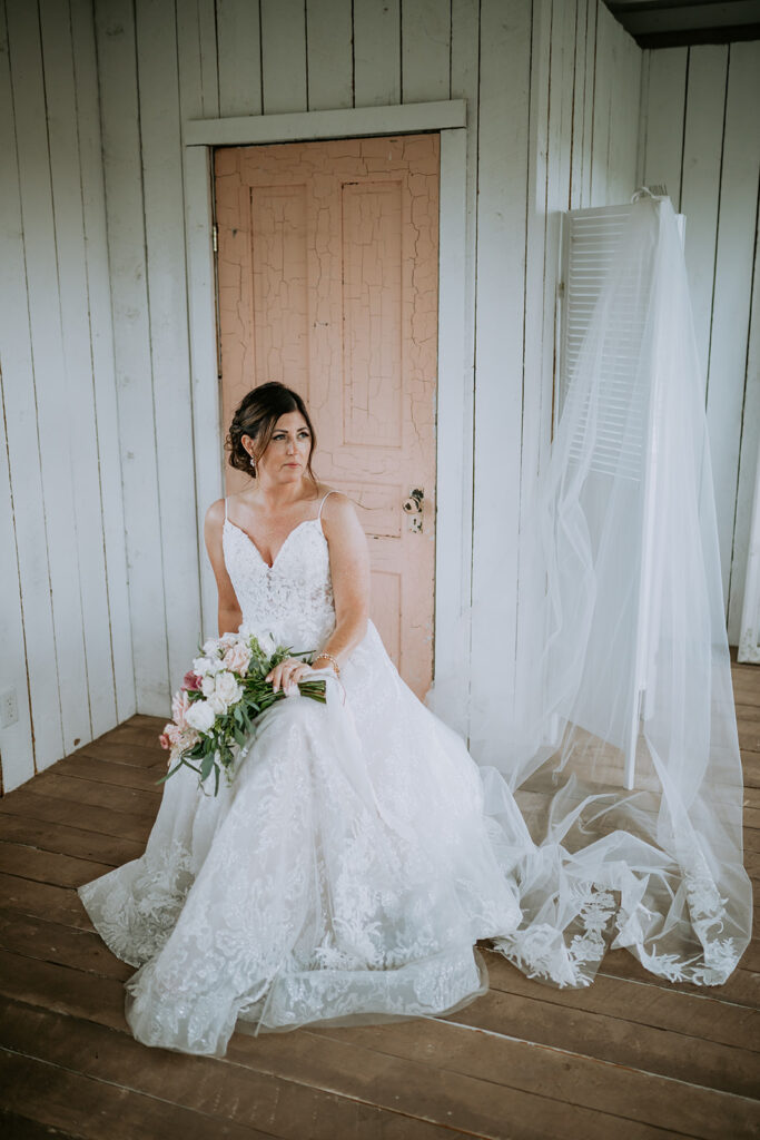 A bride in a lace wedding dress holding a bouquet sits contemplatively near a door with her veil draped beside her.