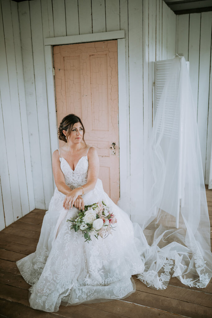 A bride sitting thoughtfully in a white room, holding a bouquet, with her veil hanging nearby.