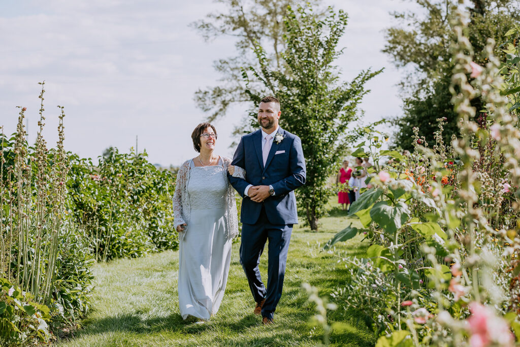 A couple walks hand in hand through a garden on a sunny day, dressed in formal wedding attire, with guests in the background.