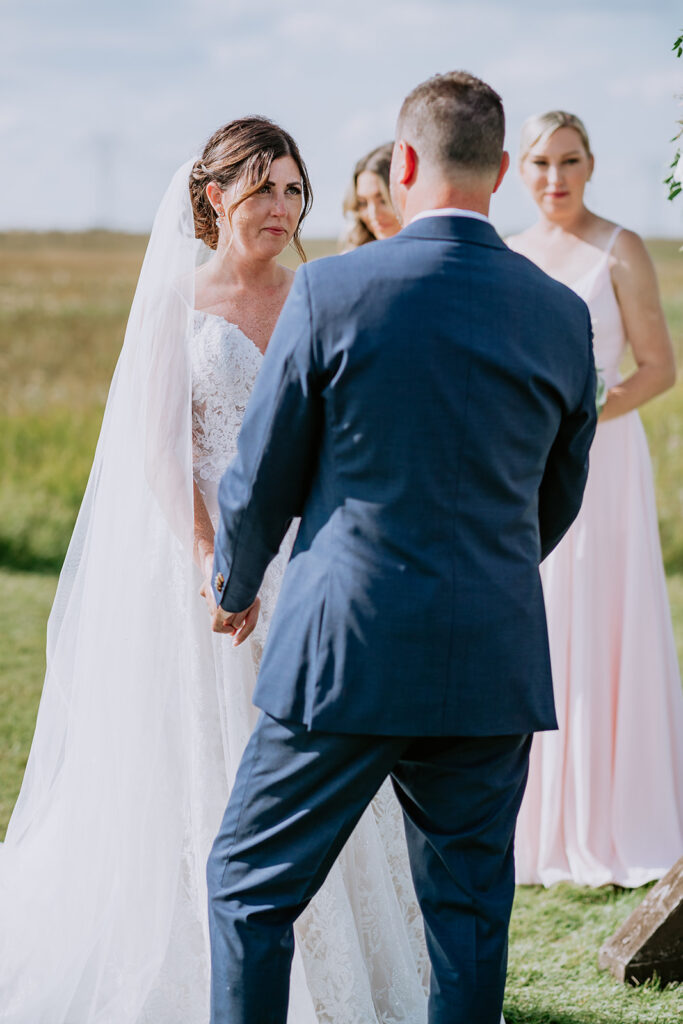 A bride holding hands with a groom outdoors, with bridesmaids watching in the background.