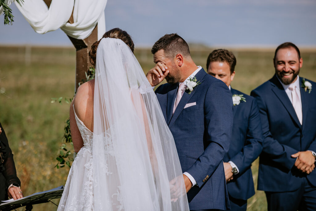 Groom wiping a tear during wedding ceremony while standing next to the bride outdoors.