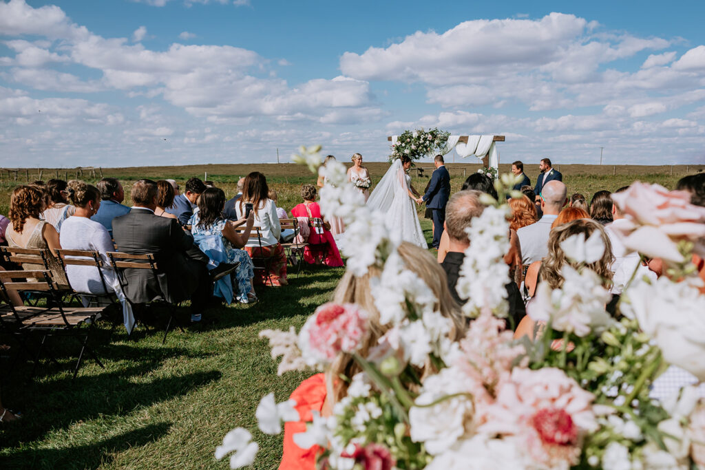 An outdoor wedding ceremony with guests seated on benches and the couple standing at the altar under a clear blue sky.